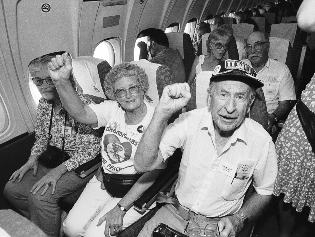 Two people giving a Spanish Civil war salute in seated inside and airplane, the woman with a grandmothers for peace t-shirt and the man with an ILWU hat.